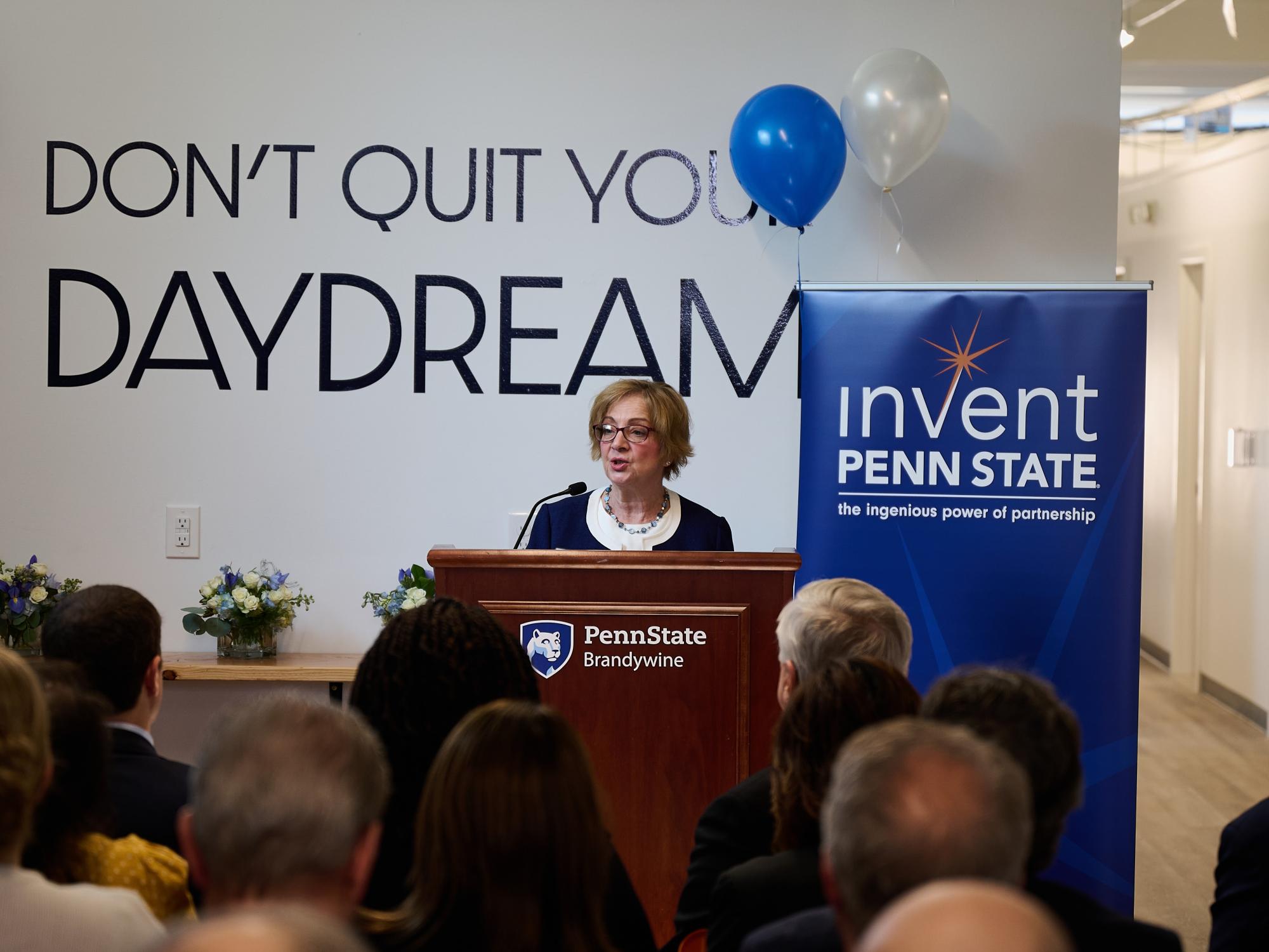 A woman stands at a lectern speaking to a crowd.