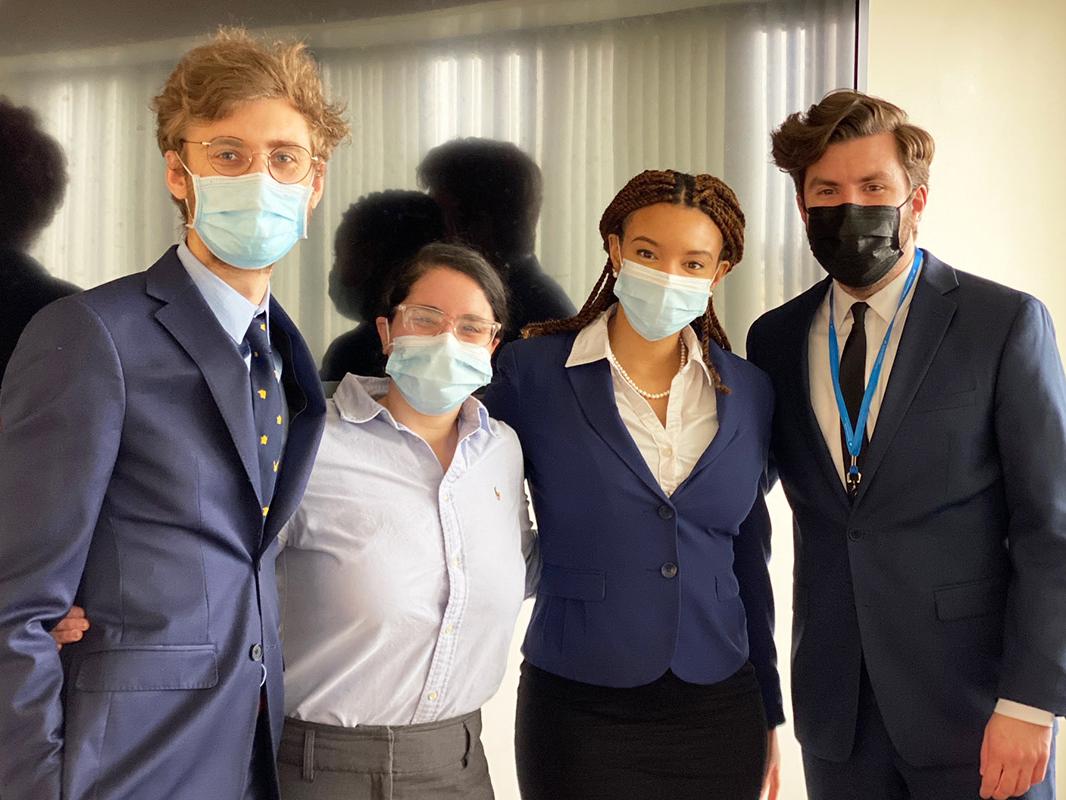 Four people stand arm in arm, wearing professional attire and masks, posing for a photo. A long mirror is on the wall in the background.