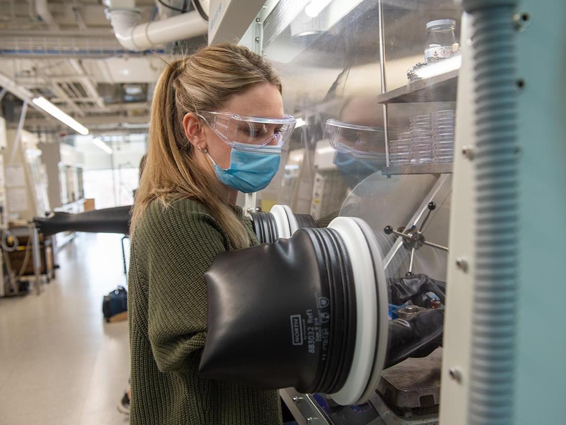 A woman stands at a machine wearing a long black glove, extending her arm into a small glass chamber in a lab.