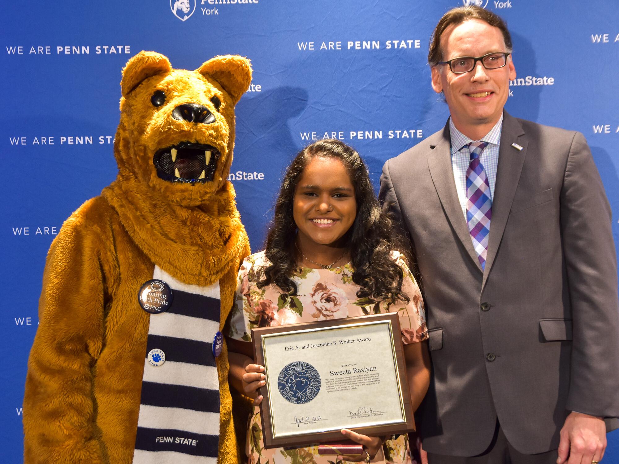 Person in Nittany Lion suit with female award recipient and male presenter at awards program.