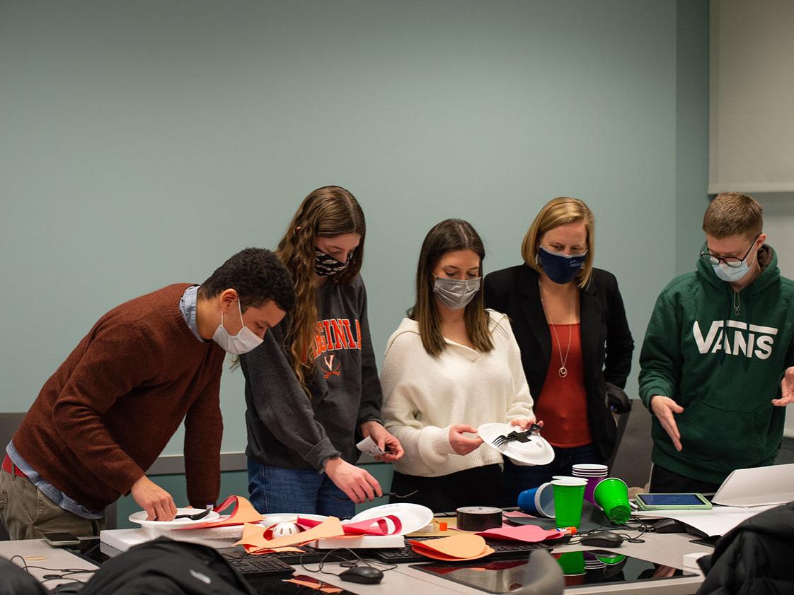 Five people wearing masks stand at a table with paper plates, plastic cups, and construction paper.