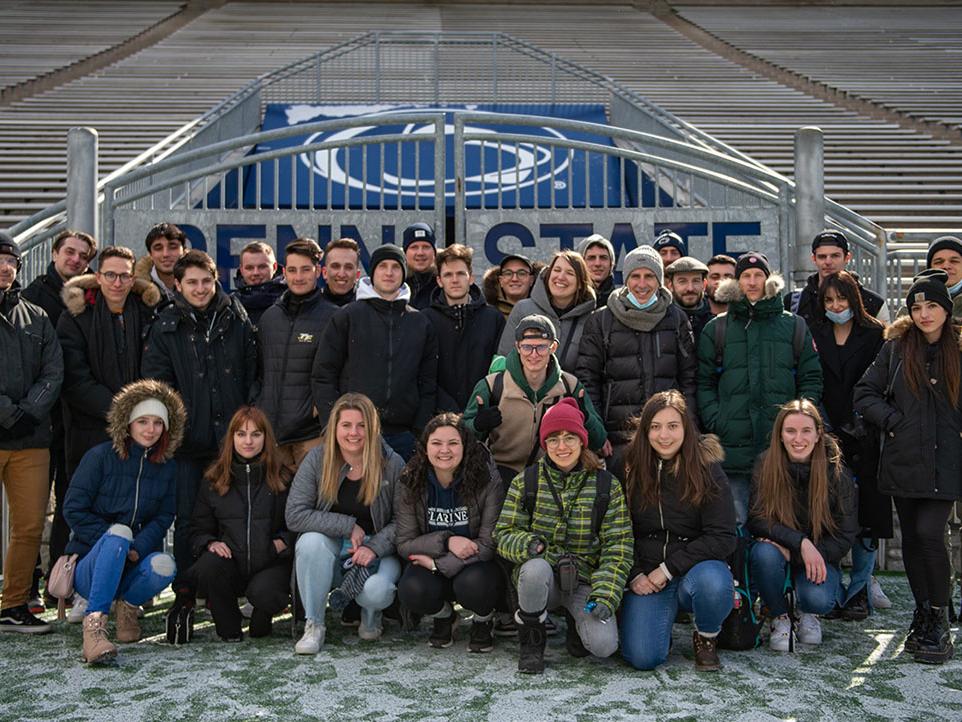 A group of people wearing winter coats poses in front of metal gates that say 'Penn State.'