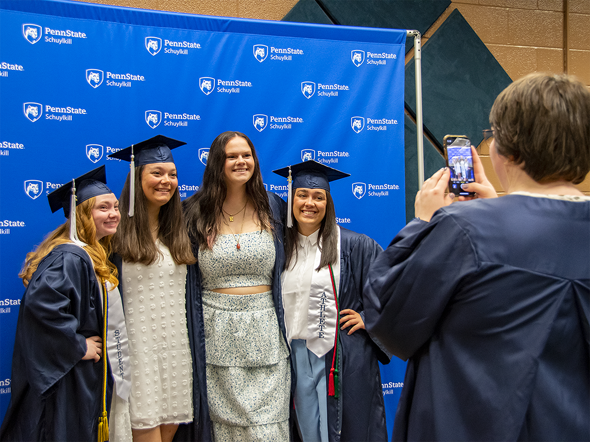 A woman takes a cell phone photo of a group of female grads against a Schuylkill branded backdrop.