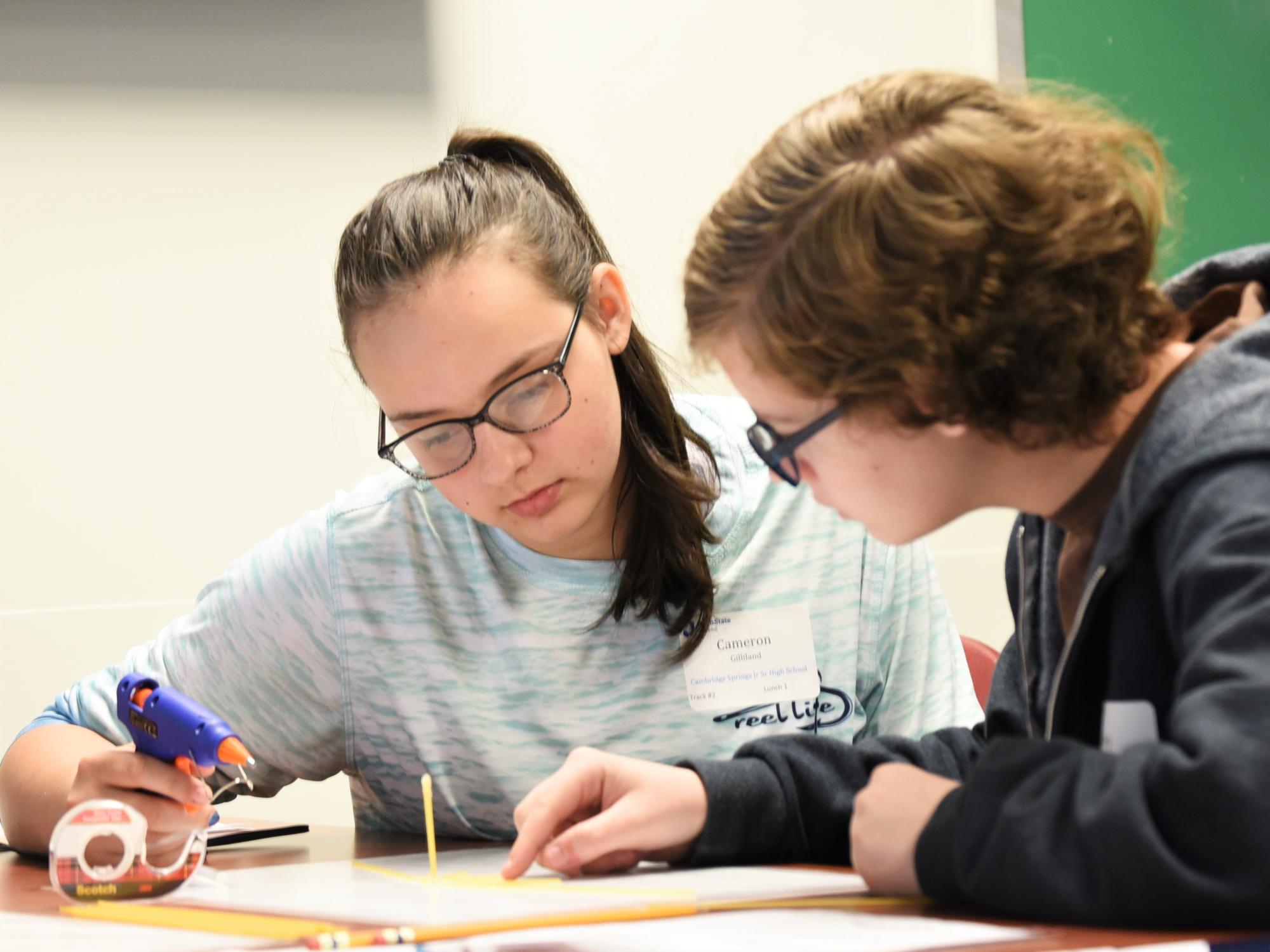 Two students build a model bridge using a glue gun and spaghetti noodles.