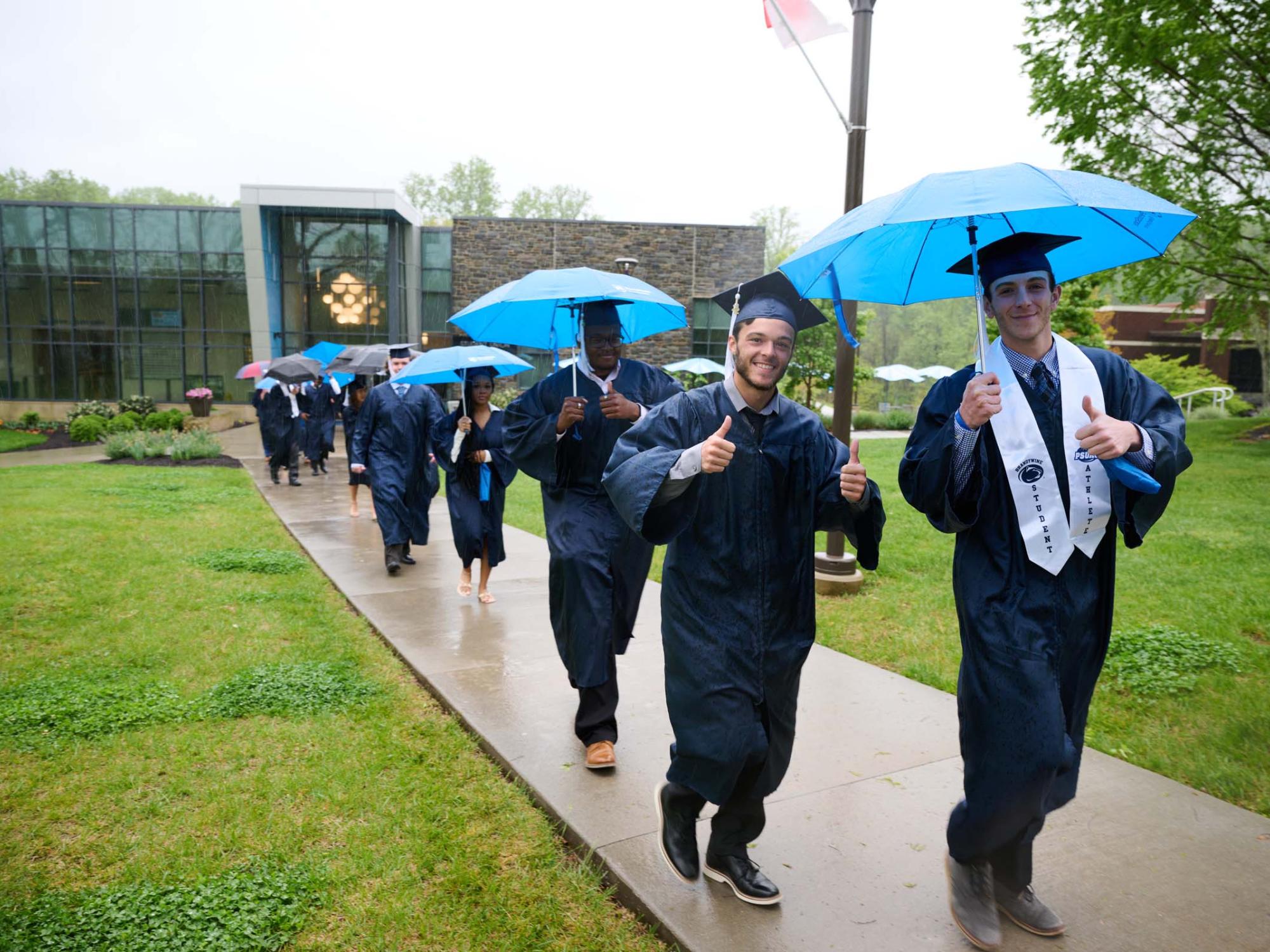 A group of students in commencement robes walks in the rain outside.