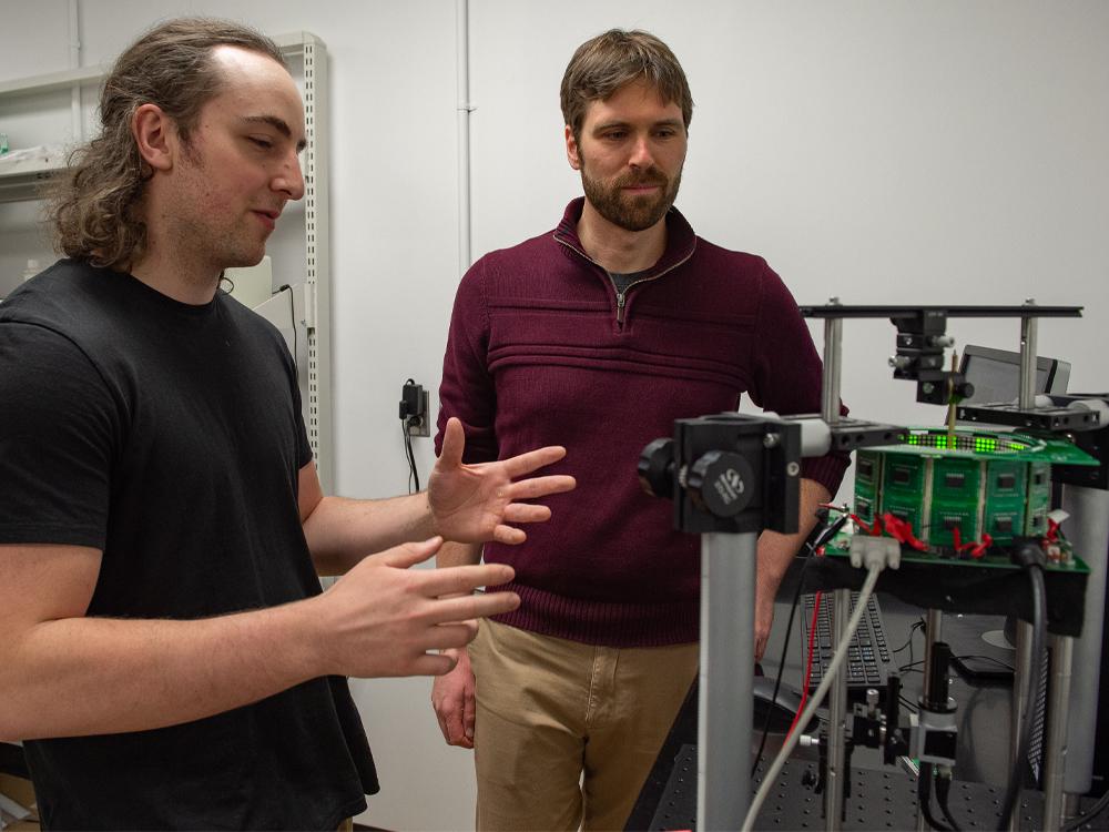Two men talk and gesture next to a piece of lab equipment.