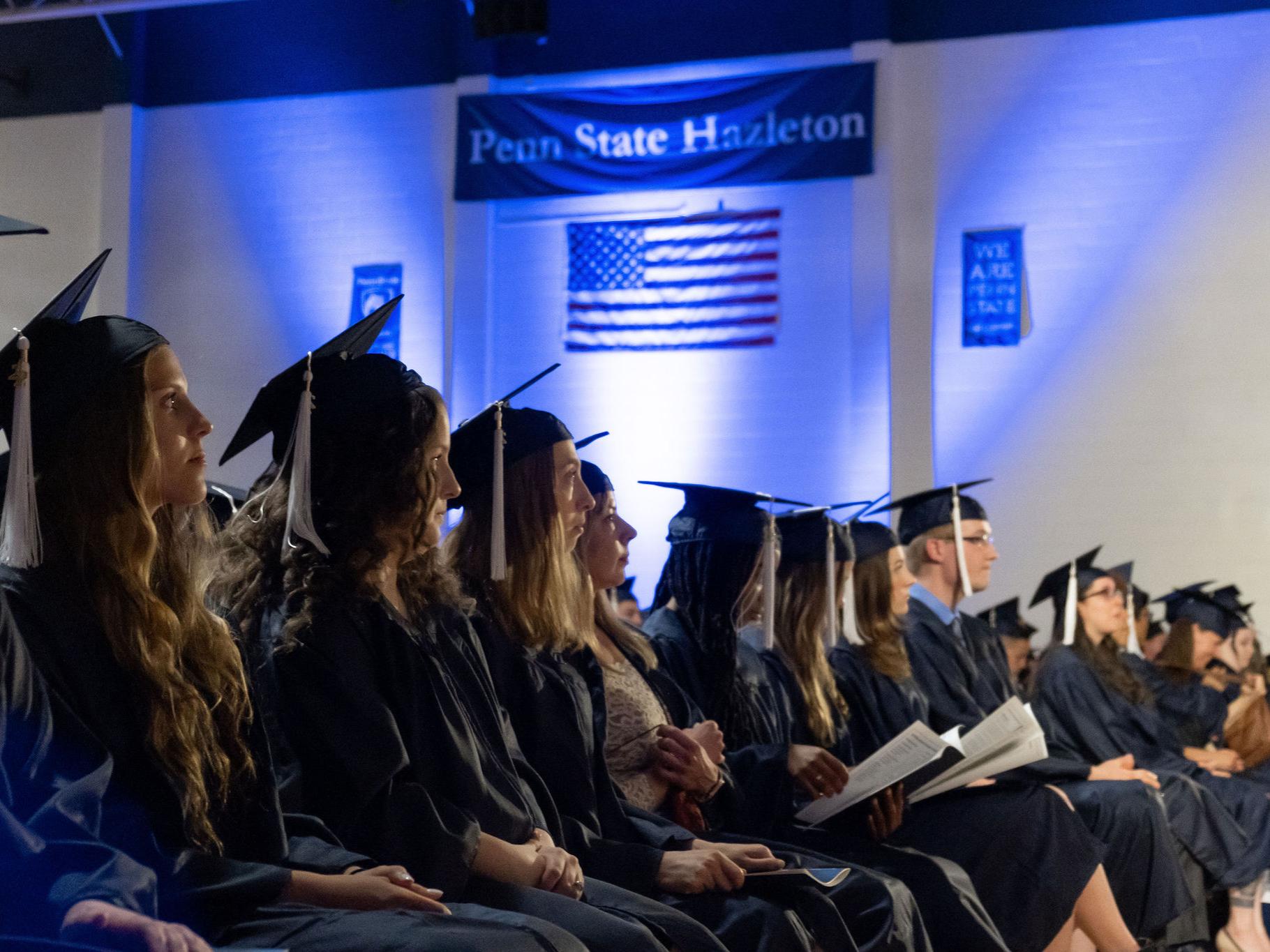 Students in caps and gowns facing a stage with Penn State Hazleton banner in background.