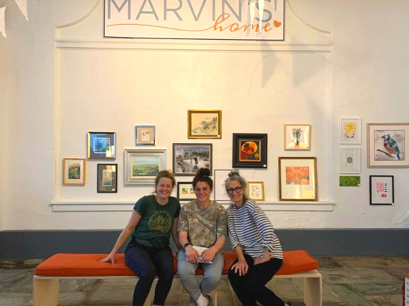 Three women sitting in front of a Marvin's Home sign and hanging artwork inside of the Marvin's Home physical storefront