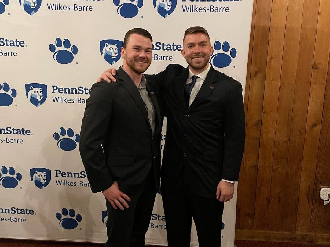 Two men in suits stand next to one another with a Penn State Wilkes-Barre backdrop in the background
