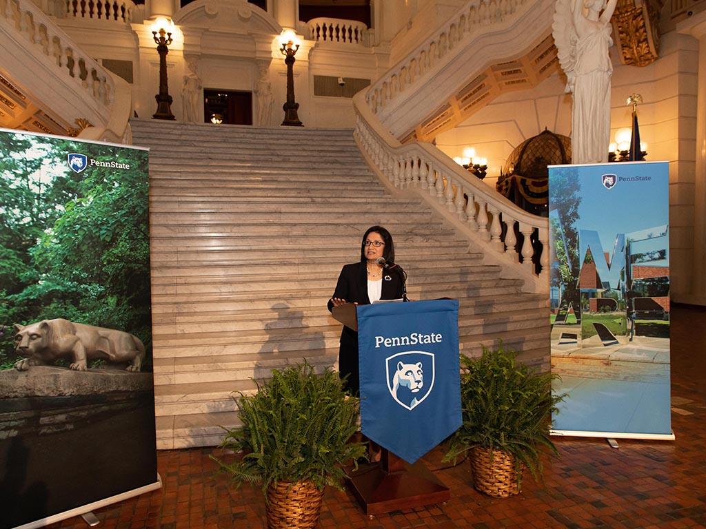 Penn State President Neeli Bendapudi speaking at a podium with a staircase behind her in the Capitol rotunda