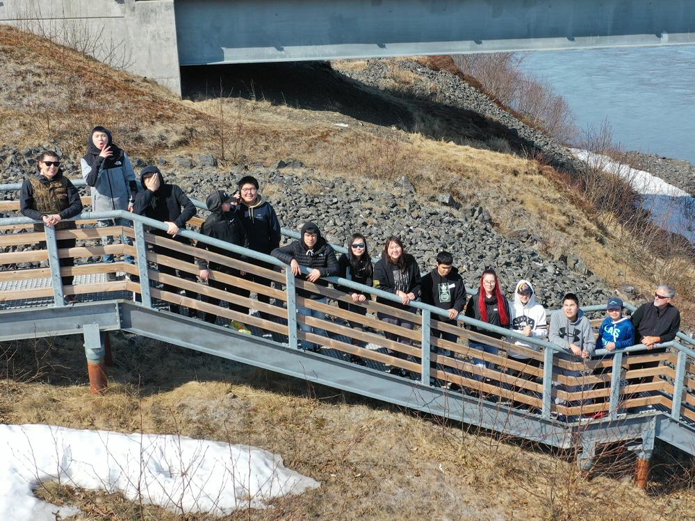14 people pose on outdoor stairs near a river