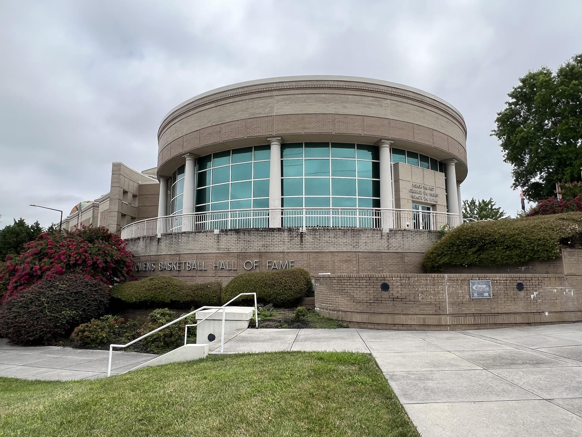 A circular building with big glass windows and manicured landscaping with a sign reading "Women's Basketball Hall of Fame"