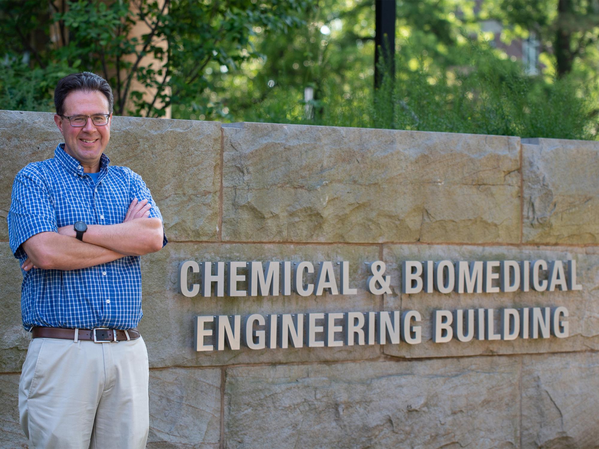 A man standing outside smiles at the camera. Behind him is a sign reading "Chemical Engineering and Biomedical Engineering Building."