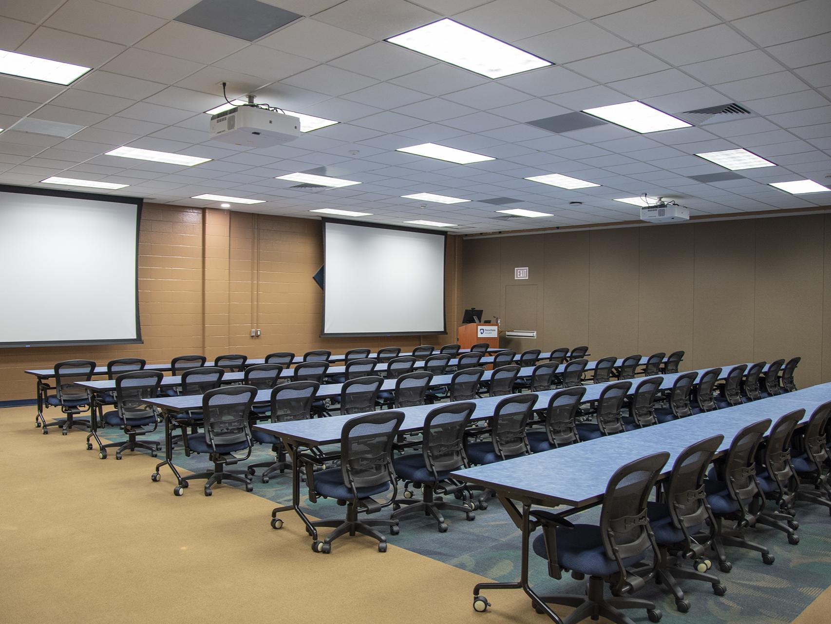 Room with brown walls and two projector screens hanging from drop ceiling with long blue tables and black chairs on wheels