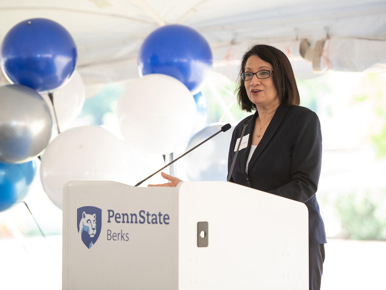 Penn State President Neeli Bendapudi speaks at a podium displaying the Penn State Berks mark, with blue and white balloons in the background. 