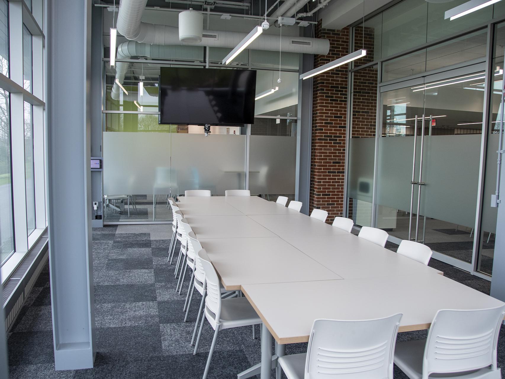 Room with floor-to-ceiling glass windows, steel support beams, and long white tables with white chairs overlooking a large monitor screen