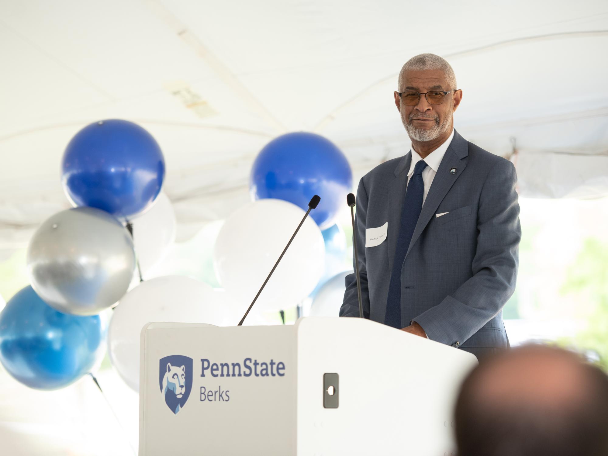 Penn State Berks Chancellor George Grant Jr. standing a podium with the Penn State Berks mark and blue and white balloons in the background.