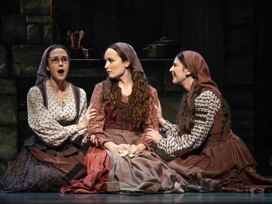 Three young ladies sit on the ground in their dresses and sing to each other.