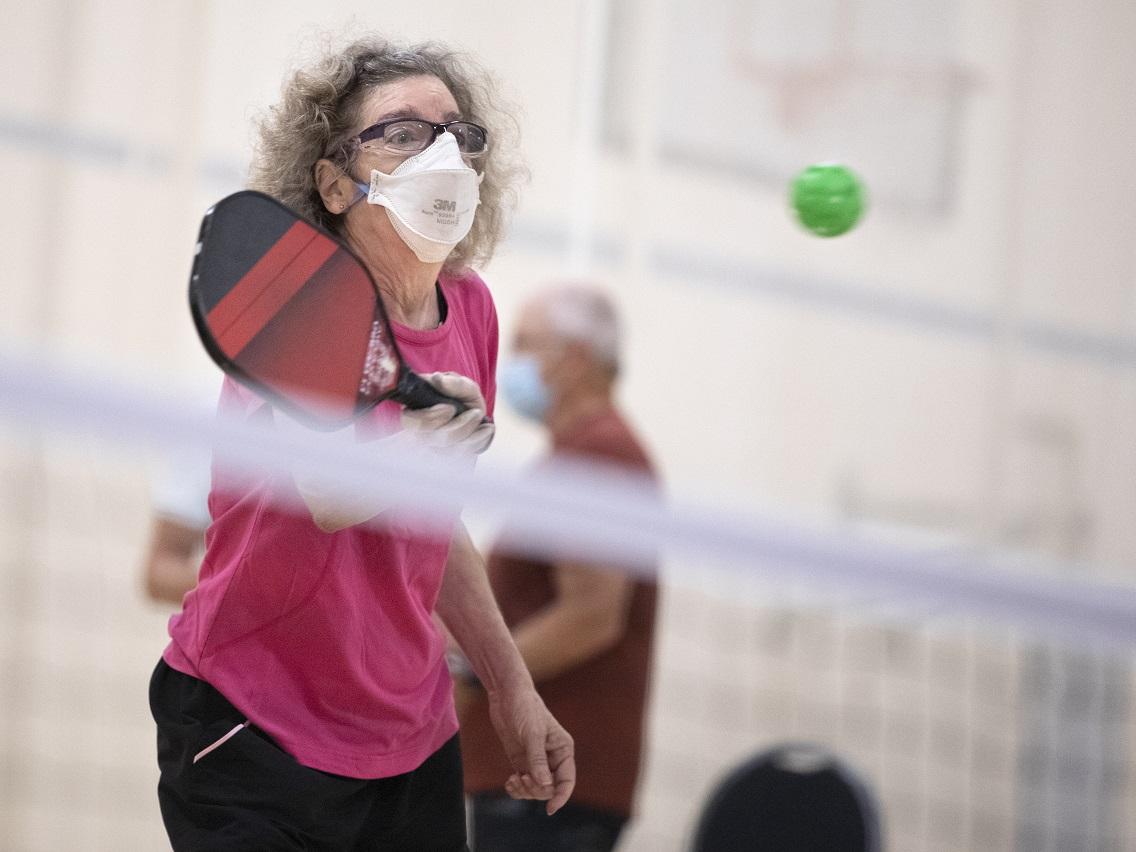 A man in surgical mask charges a net brandishing a pickleball racquet in a gym. Two other men are seen from behind in the foreground.