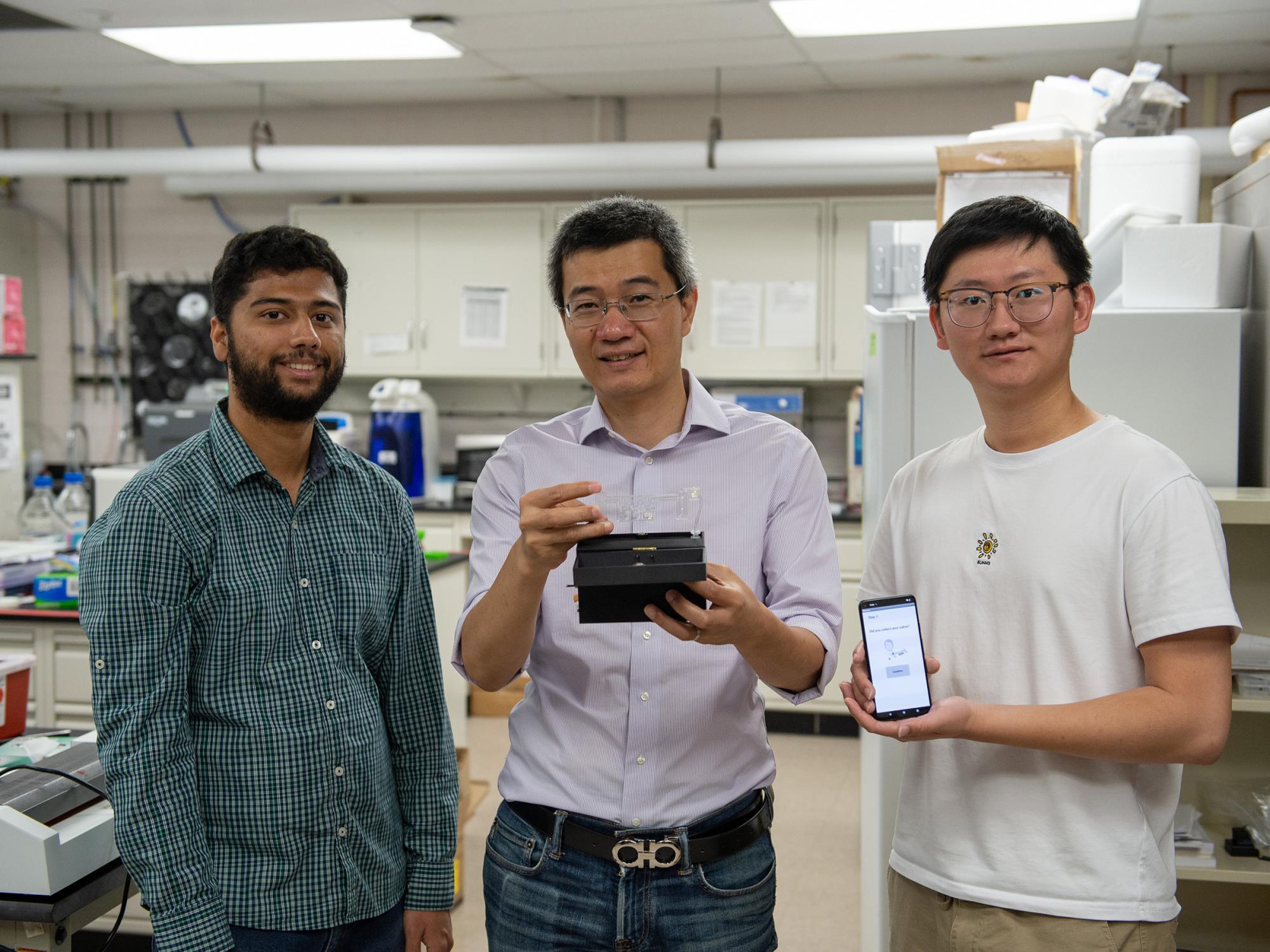 Three individuals pose in a lab holding a COVID-19 test prototype and a smartphone. 
