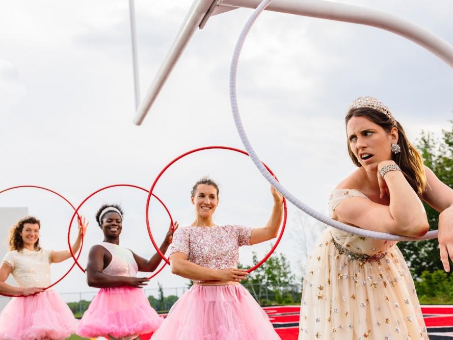 Four women wearing tutus, one with a scowl, hold Hula Hoops while standing near a goalpost.
