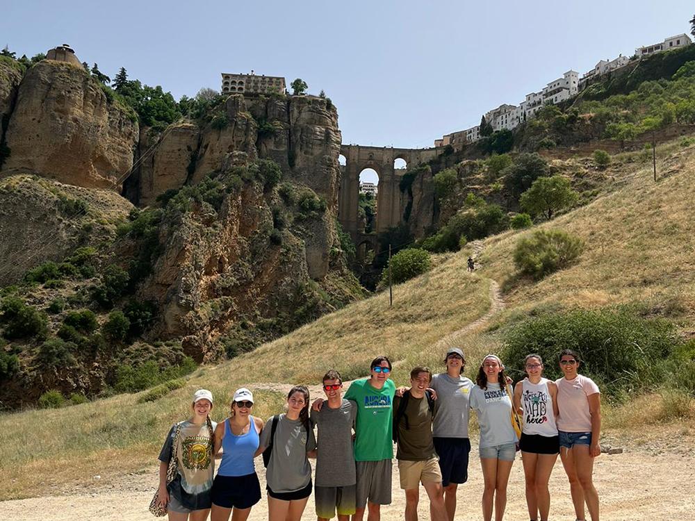Ten college students stand in the foreground with Centro de Interpretación del Puente Nuevo in Ronda, Spain in the distance behind them