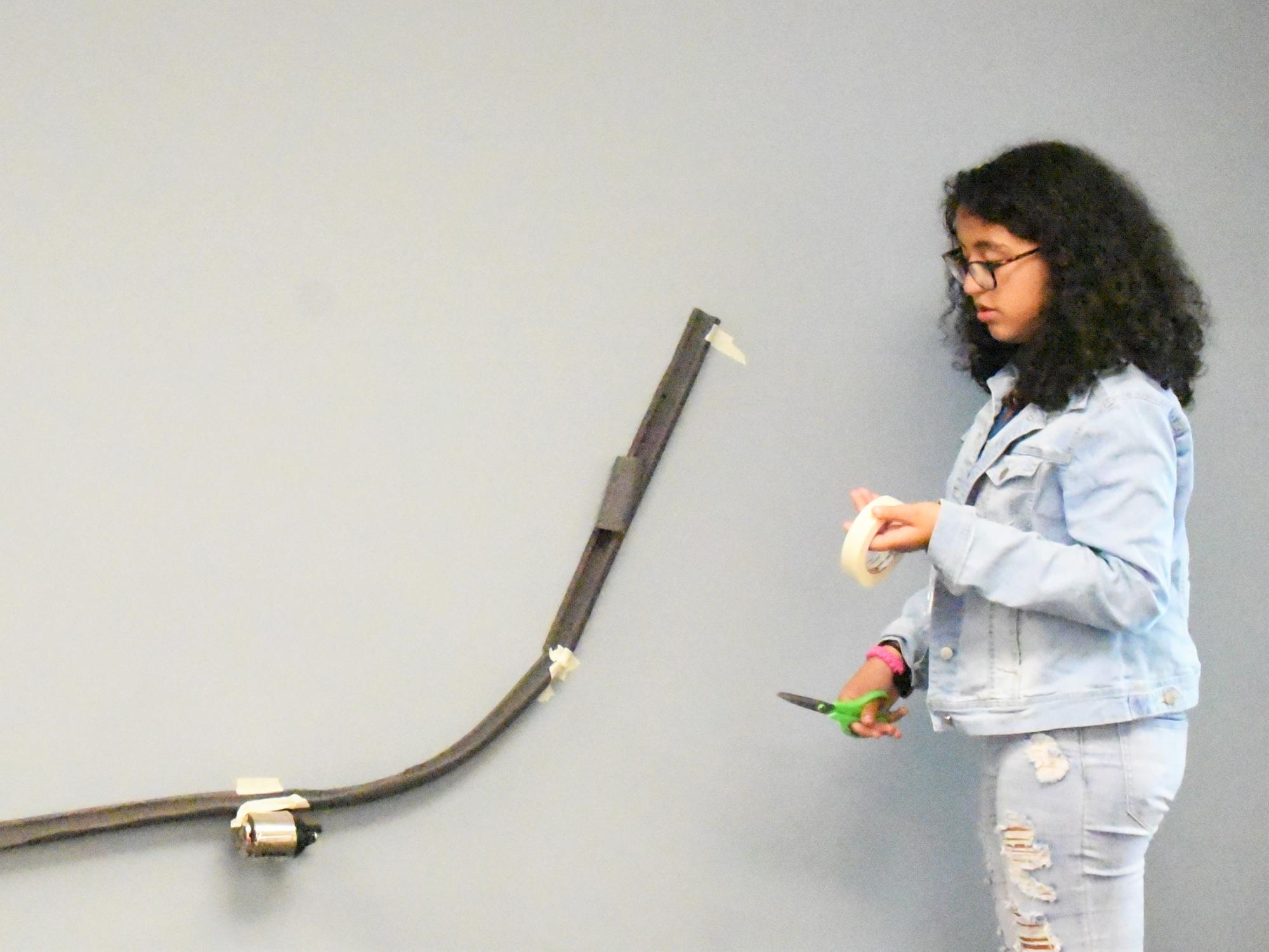 A girl holding tape and scissors stands next to a model roller coaster track, which is hung on the wall with pipe insulators.