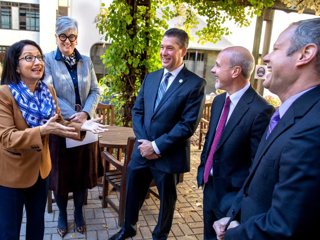 Five people stand on a brick patio near a leaf-covered terrace, talking and smiling.