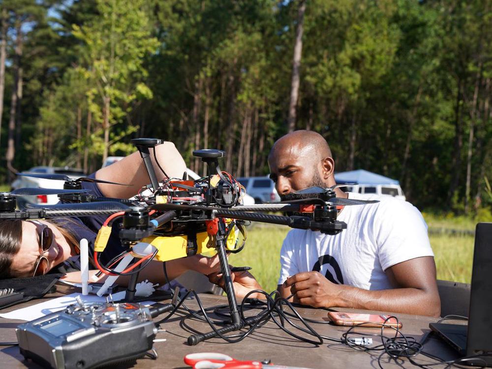 Two individuals adjust a drone on a table. 
