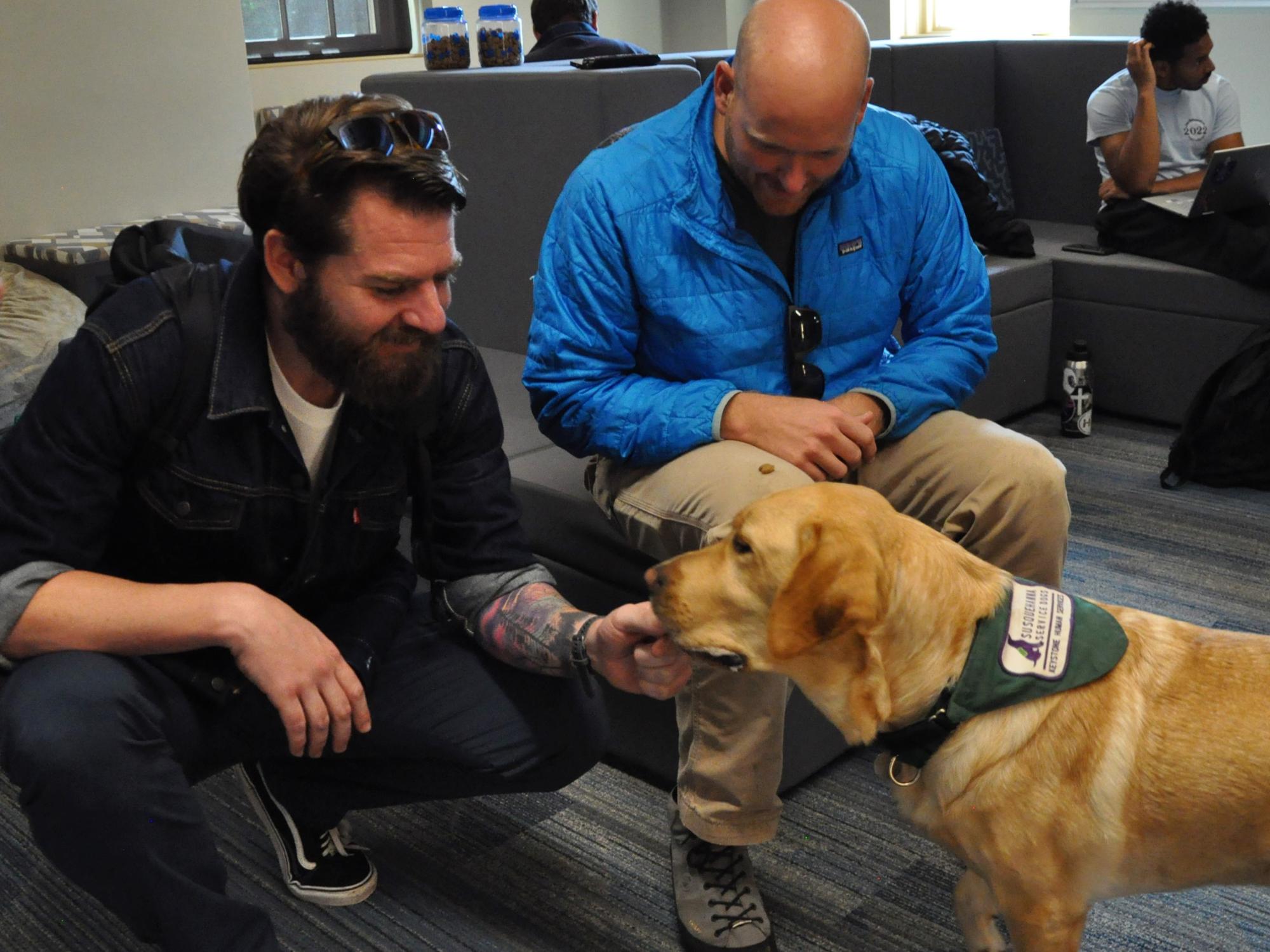 Deux étudiants seniors de Penn State sont assis avec le chien de l'établissement, Podrick, souriant et se grattant le menton.