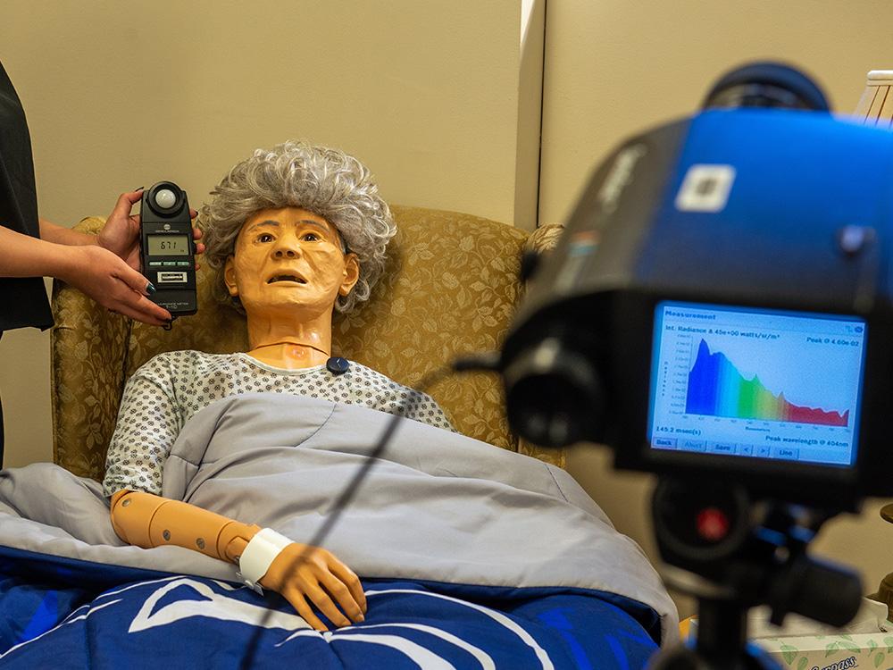 Dummy of elderly woman wearing a monitor and surrounded by equipment is used for light experiment.