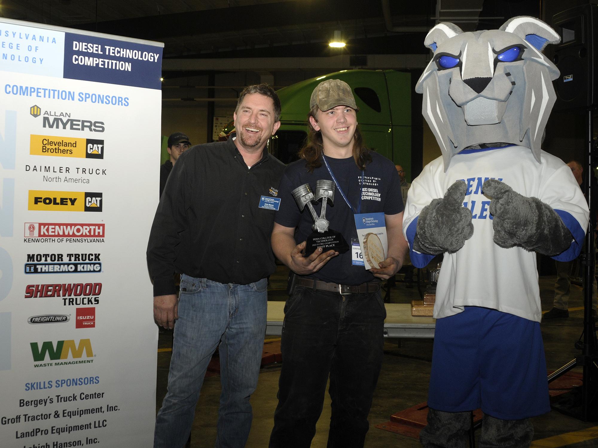 Two people and the Penn College Wildcat mascot stand with a trophy