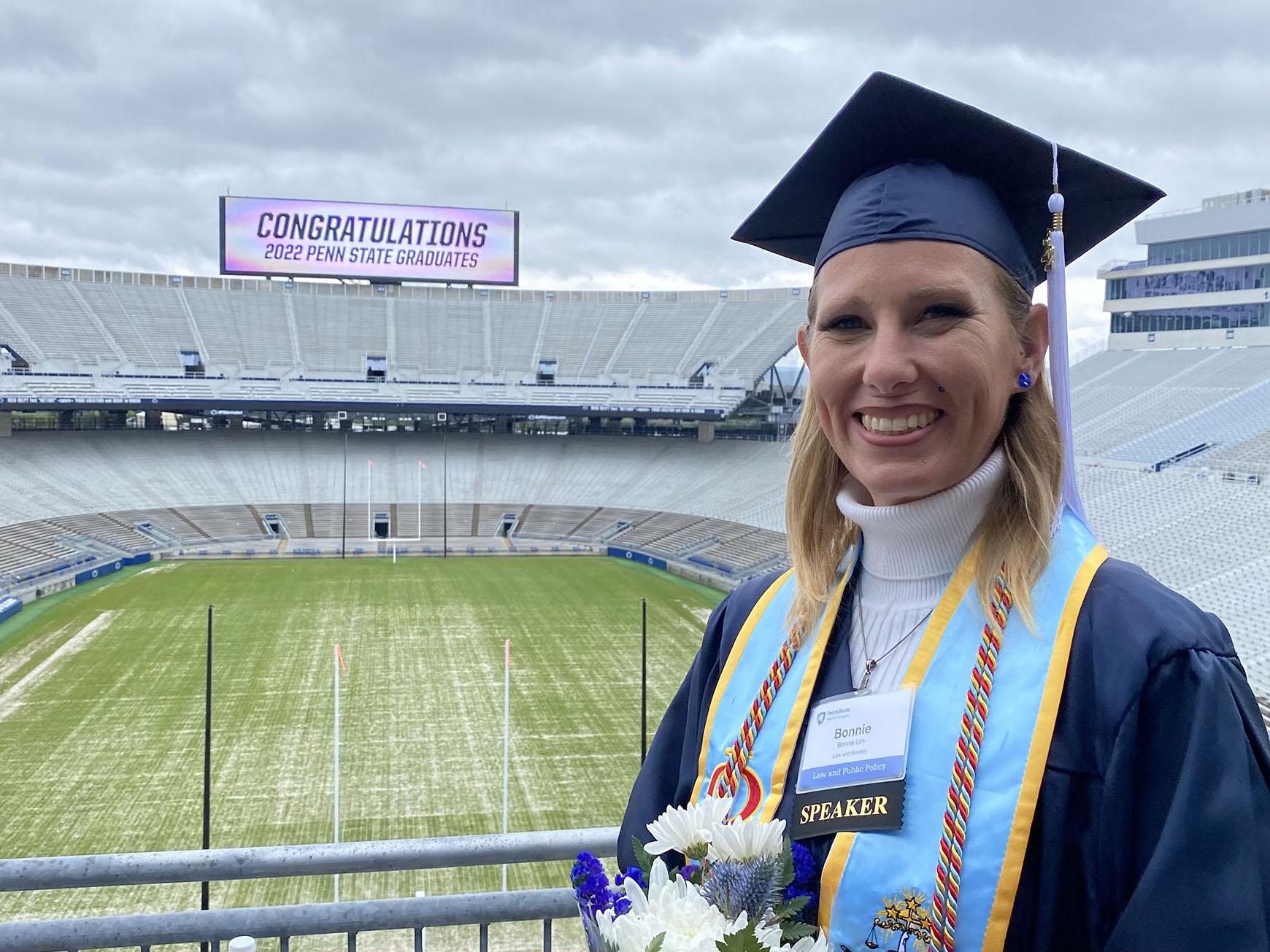 Bonnie Lim, wearing a cap and gown, stands with the field and stands of Beaver Stadium in the background.