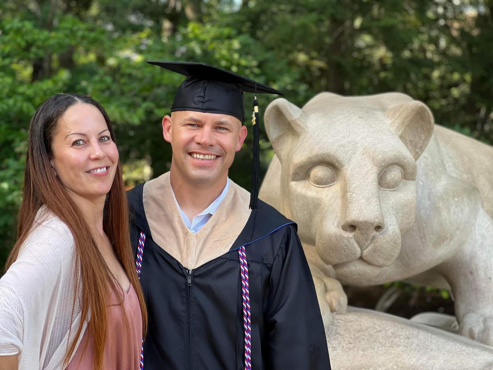 Curt Rice wears a cap, gown and military honor cords standing with a woman in front of the Nittany Lion statue.