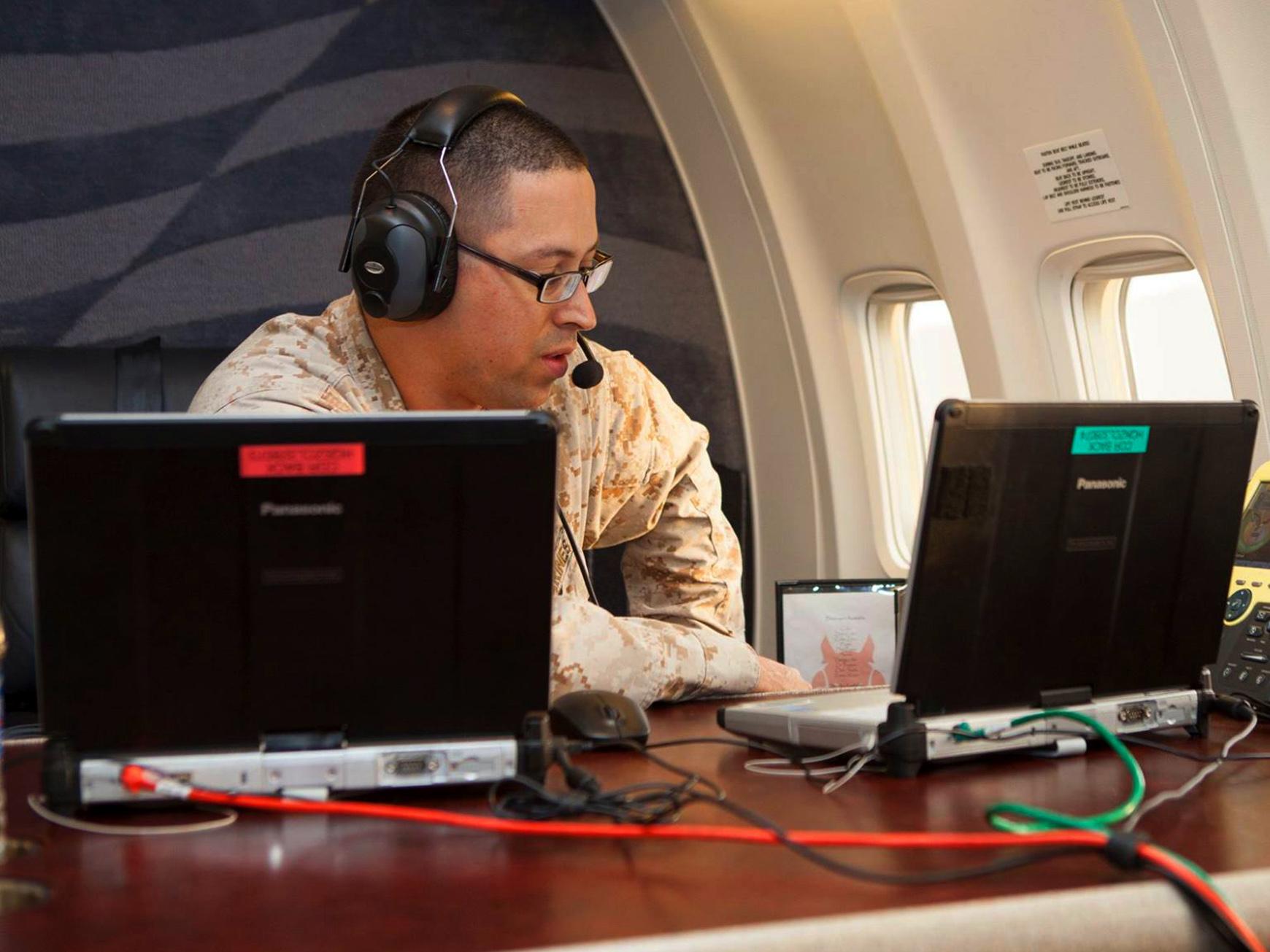 Roberto Rodriguez, wearing headphones and a microphone, works facing two computers on a military plane.