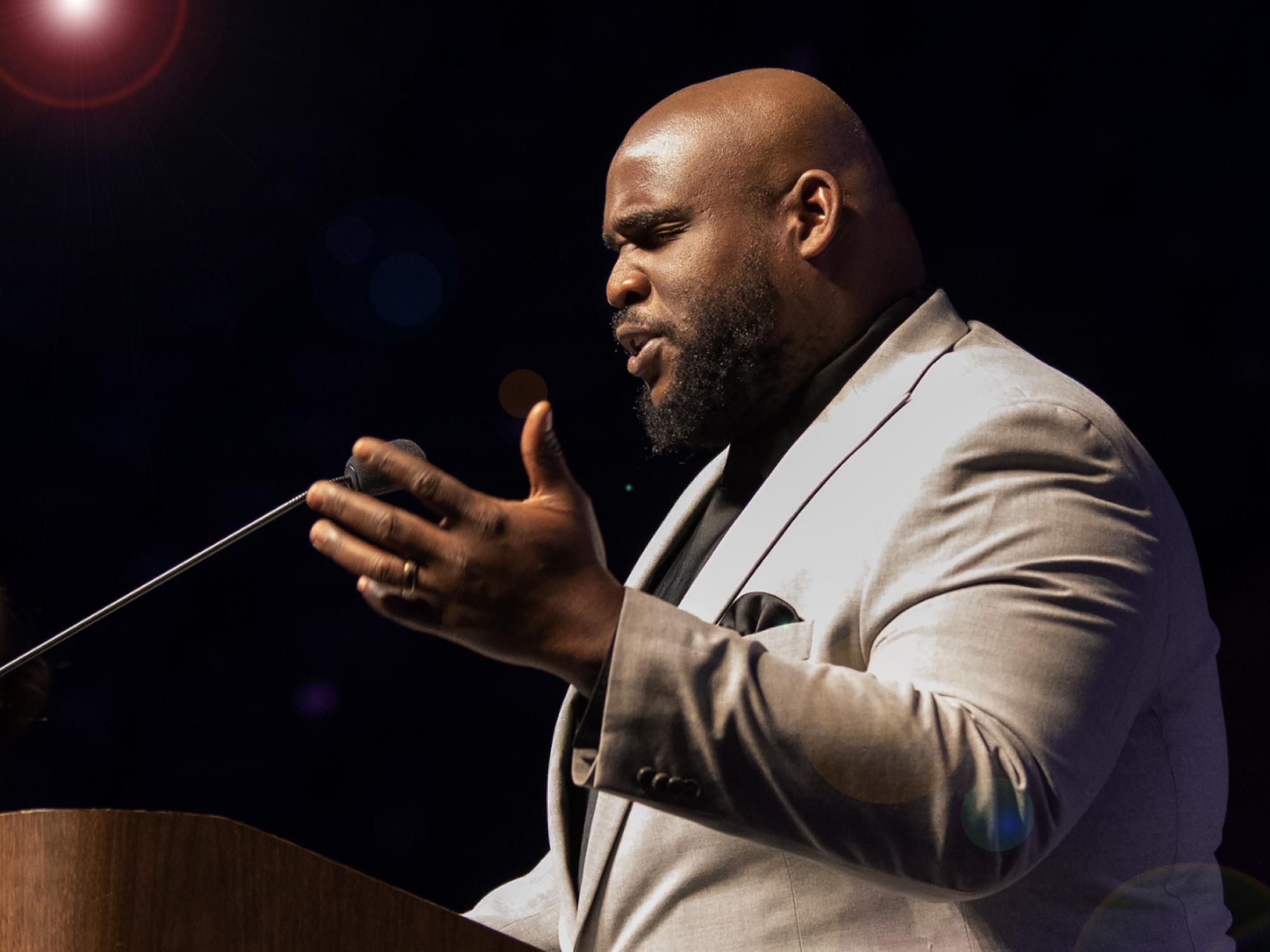Rev. Paul McReynolds, dressed in a tan suit, speaks at a podium at an event