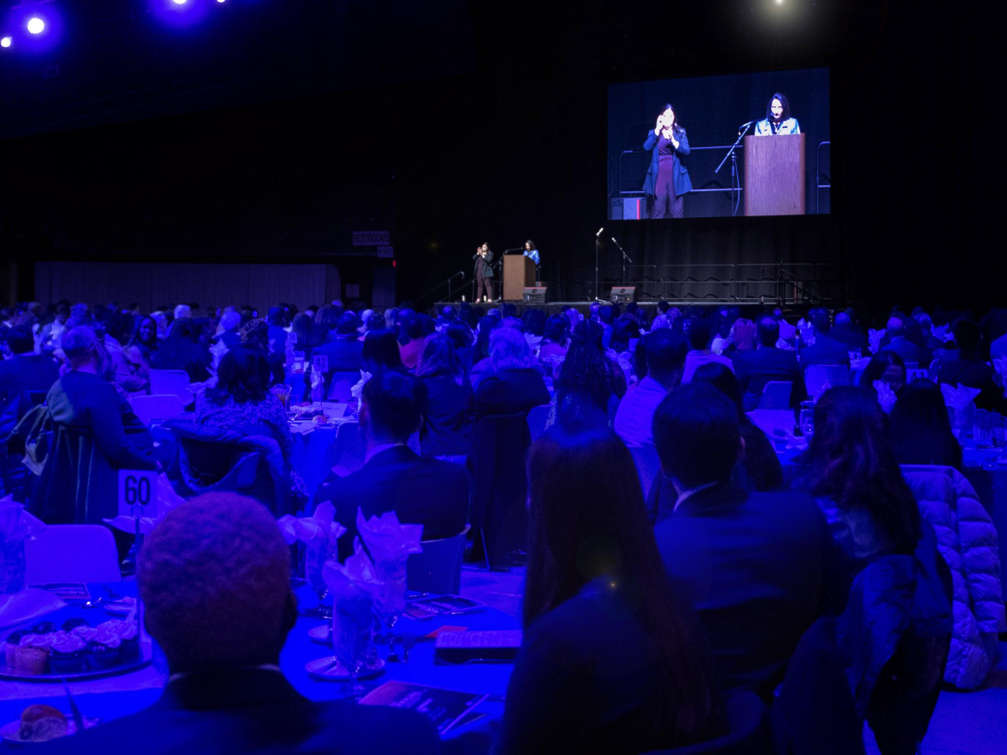 An audience seated at tables are dimly lit in blue, listening to a brightly lit speaker at a podium, with a sign language interpreter beside her.
