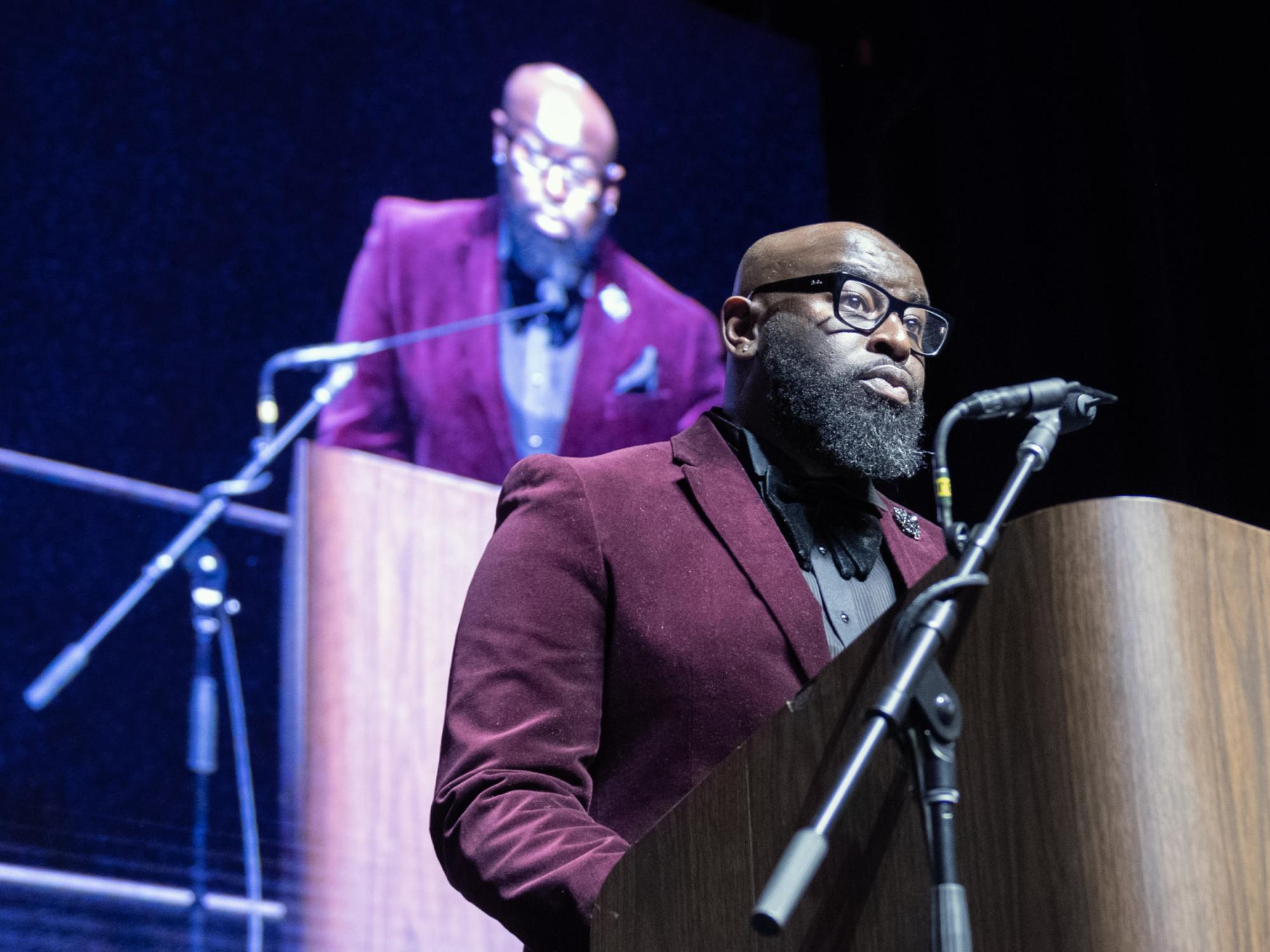 Andre Culbreath, wearing glasses and a burgundy suit jacket with bow tie, speaks at a podium.