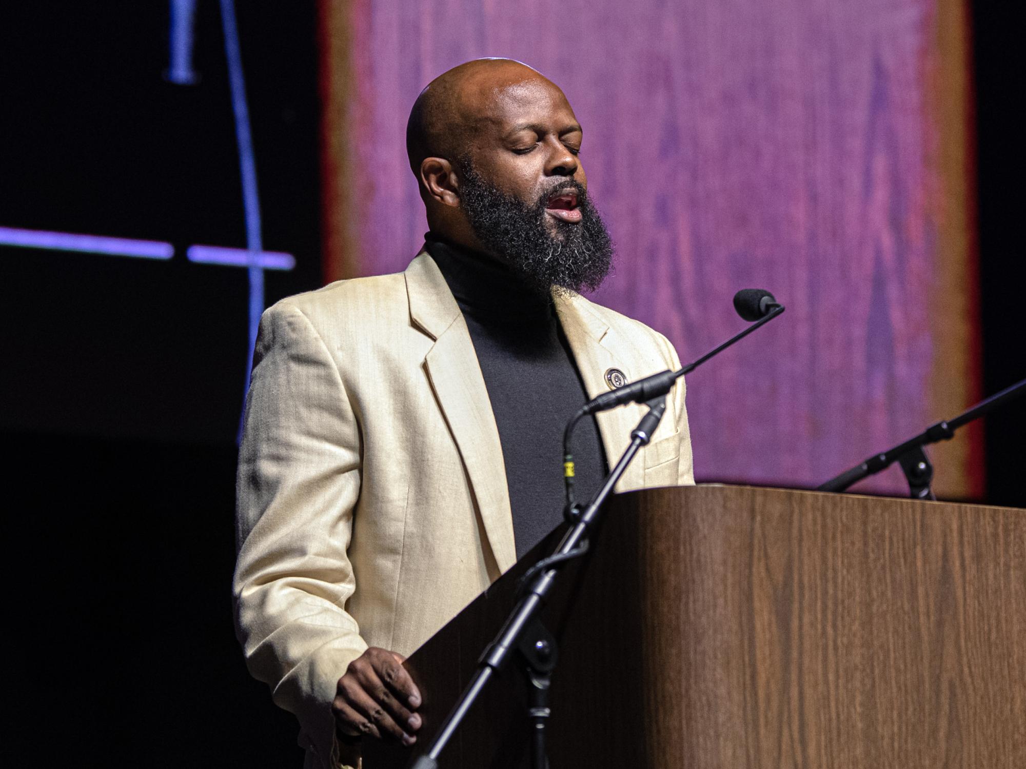 Gabriel Green, wearing a cream-colored jacket and black shirt, sings at a podium.