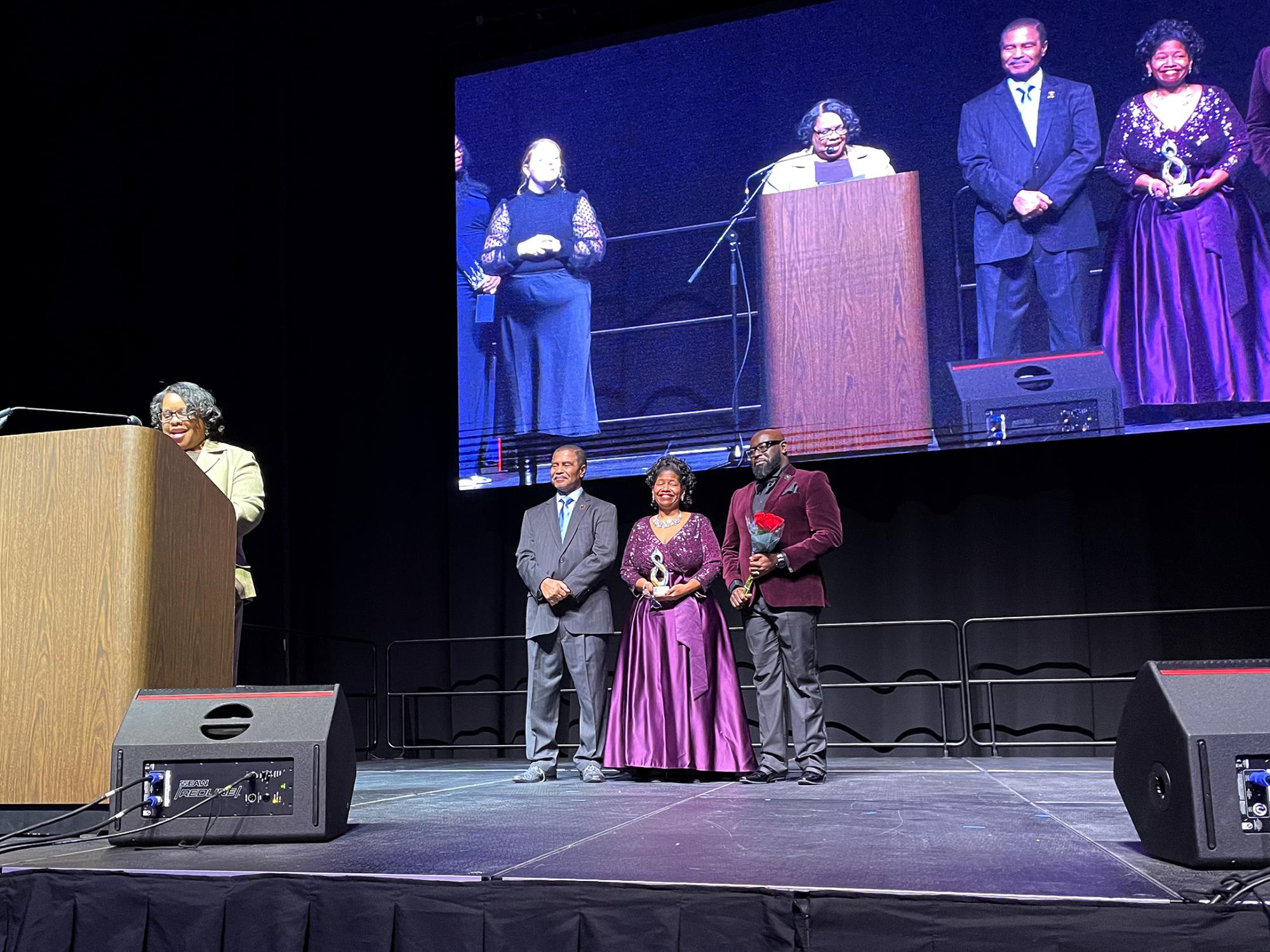 Several people in formal attire stand on a brightly lit stage at a gala event.