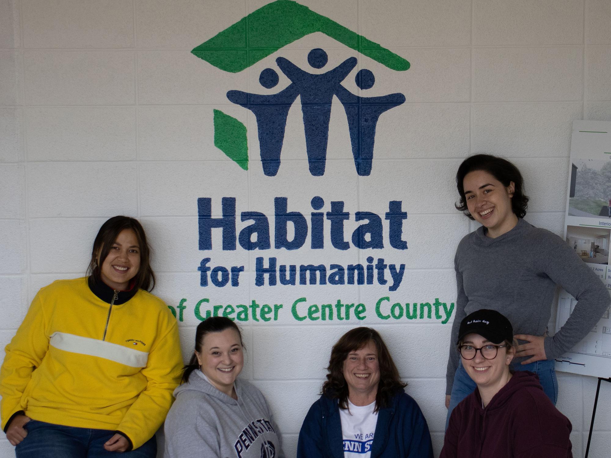 A group of five people stand in front of a white wall with a painted Habitat for Humanity logo.