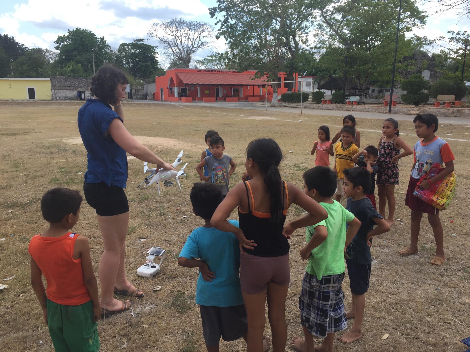Young woman holding a drone in front of small children in a field