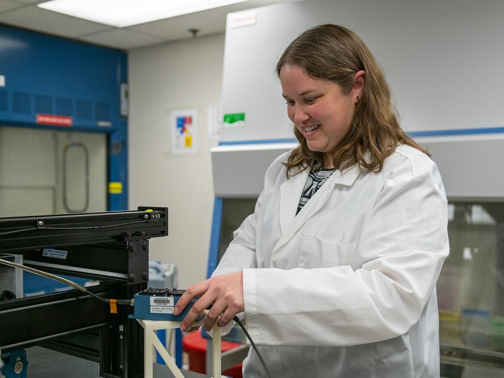 A person in a white lab coat smiles and adjusts a blue rectangular piece of equipment.
