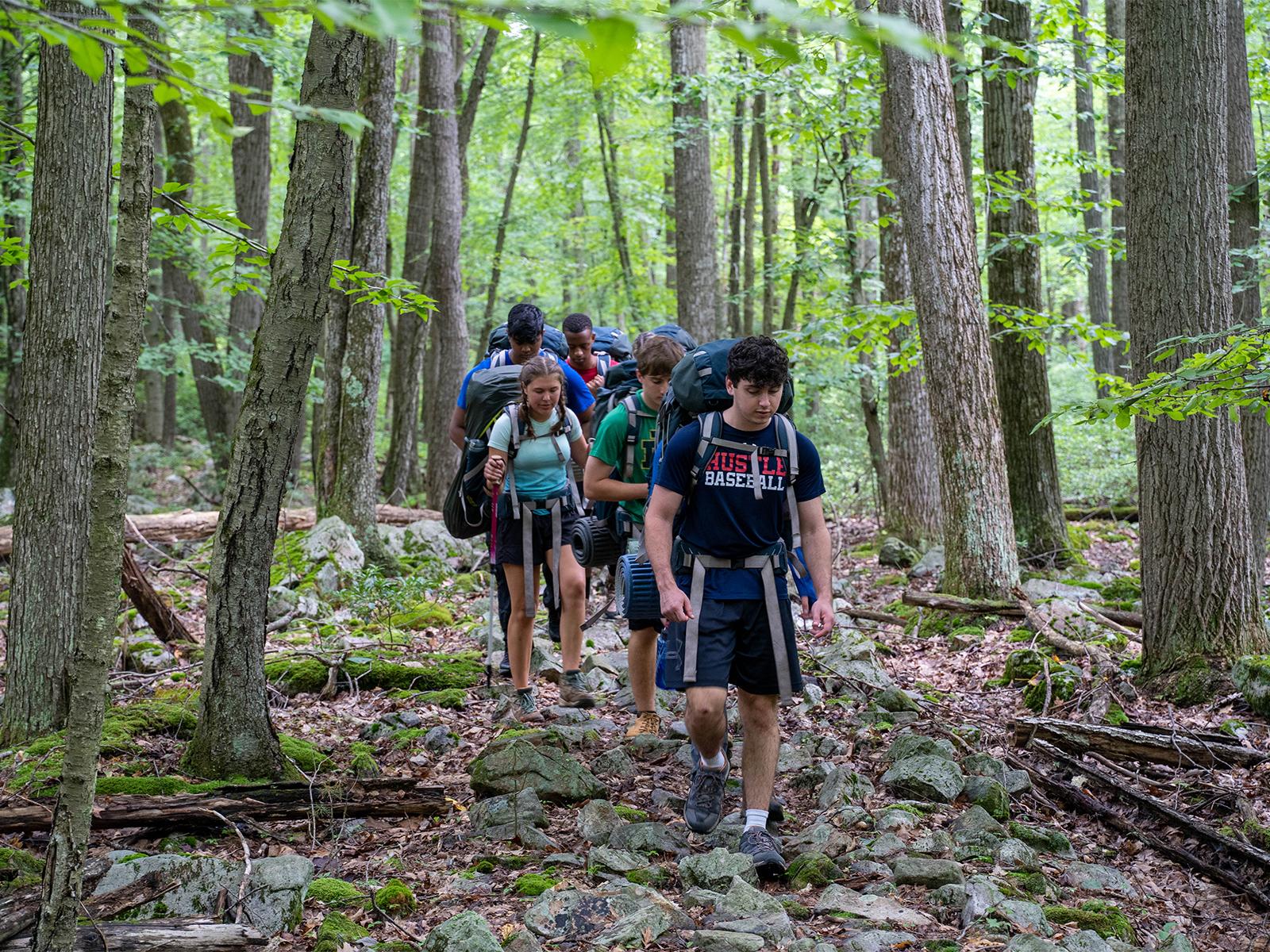 Students carrying backpacks and hiking along a trail in the woods in the summer..