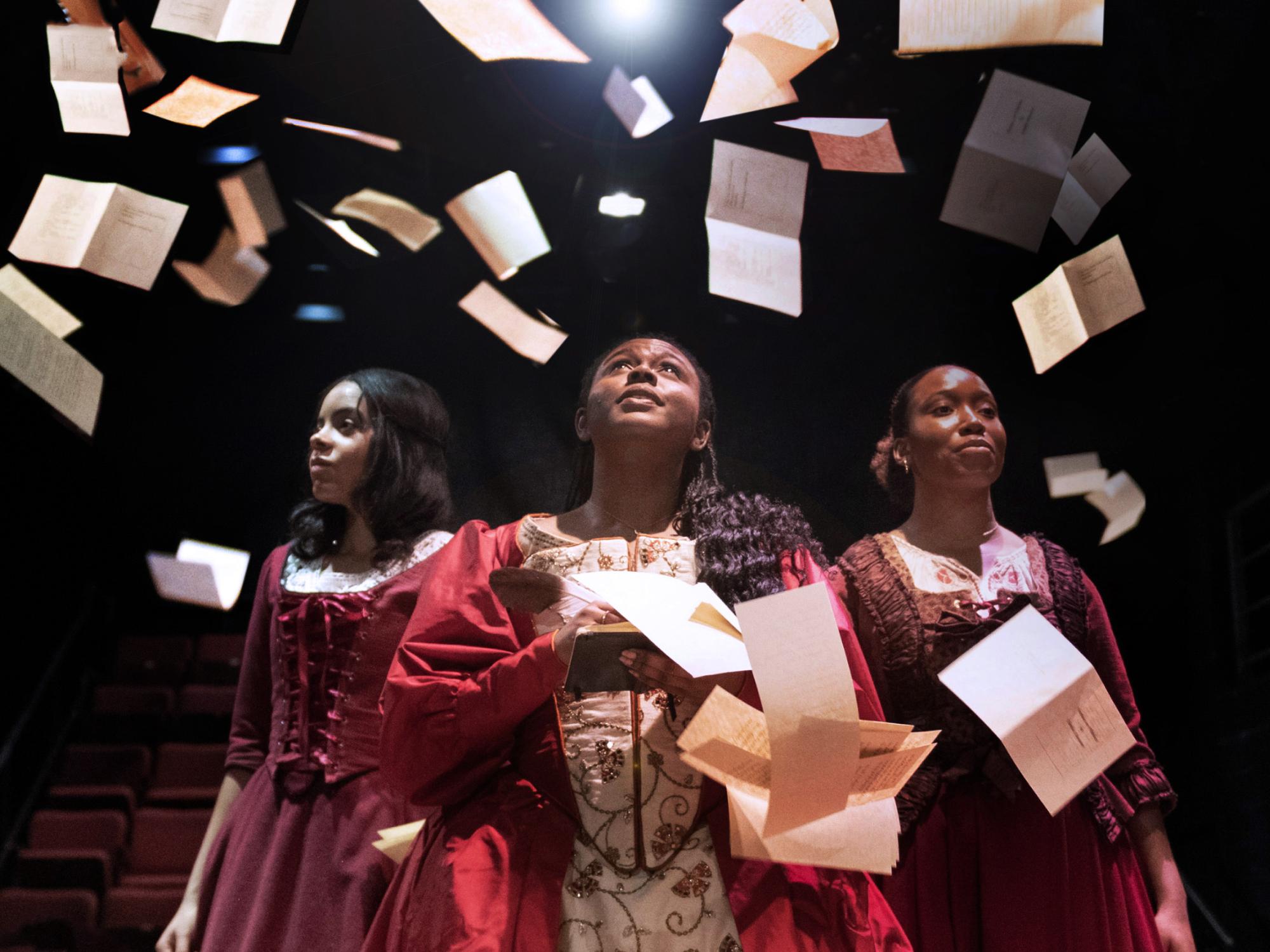 Three actors wearing seventeenth century dresses, book pages are falling all around them.
