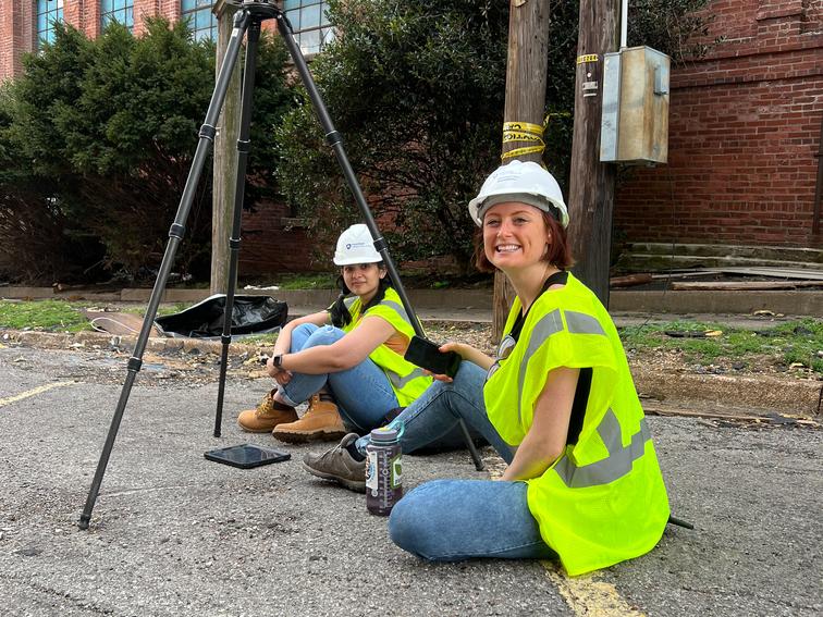 Two researchers wearing hard hats sit on the pavement following a tornado in Mayfield, Kentucky.