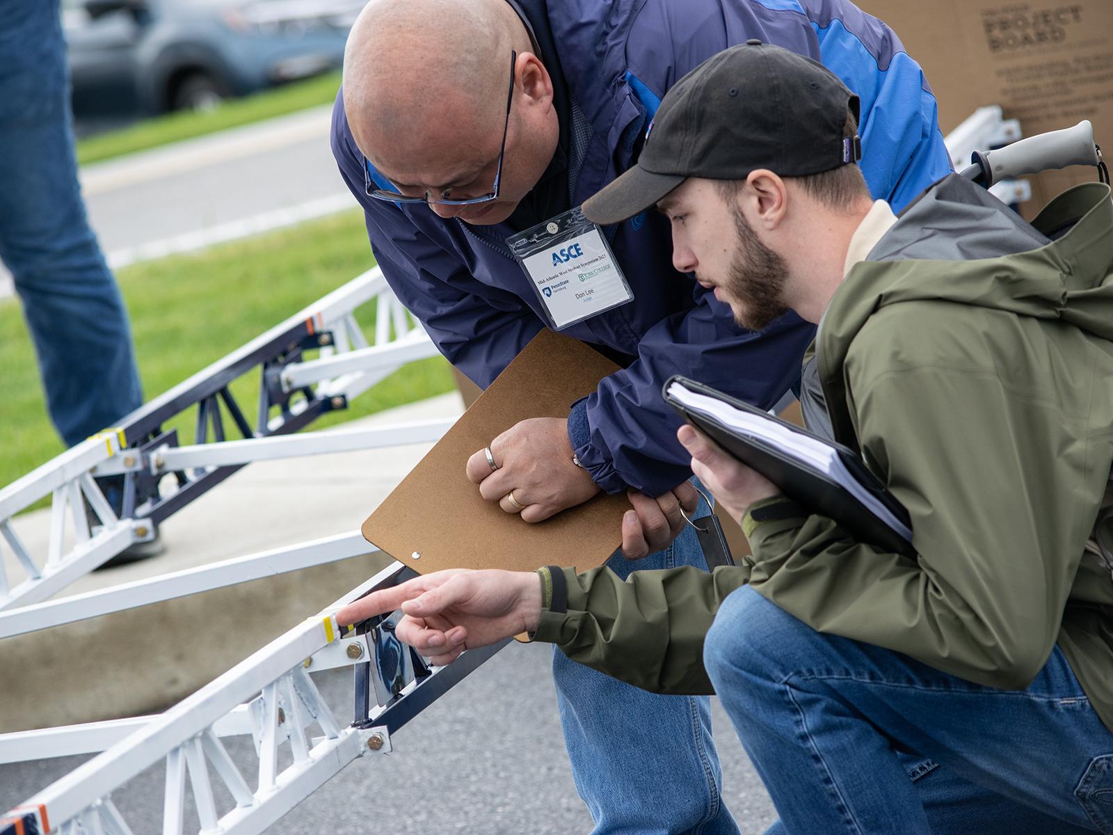 Two men holding clipboards and papers judge a steel bridge made by a student team