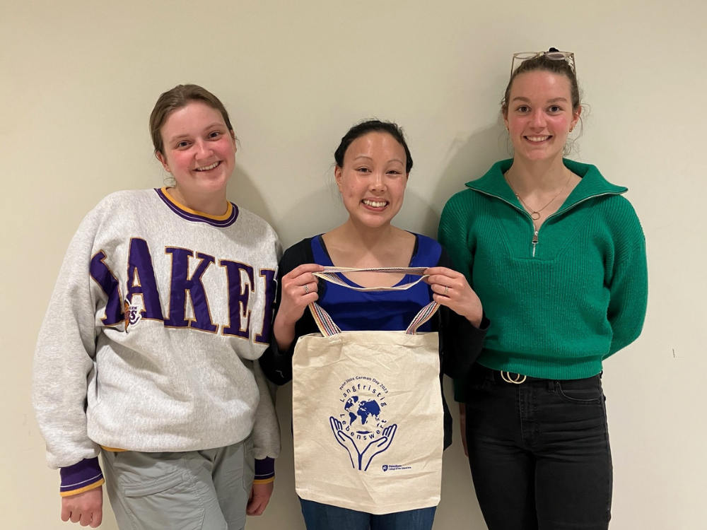 One undergraduate student and two graduate students stand in a row smiling at the camera while the student in the center holds a tote bag