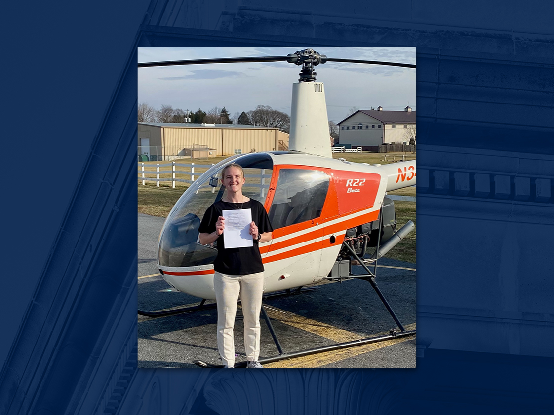 Young woman in light pants and dark shirt poses in front of orange and white helicopter on airfield.
