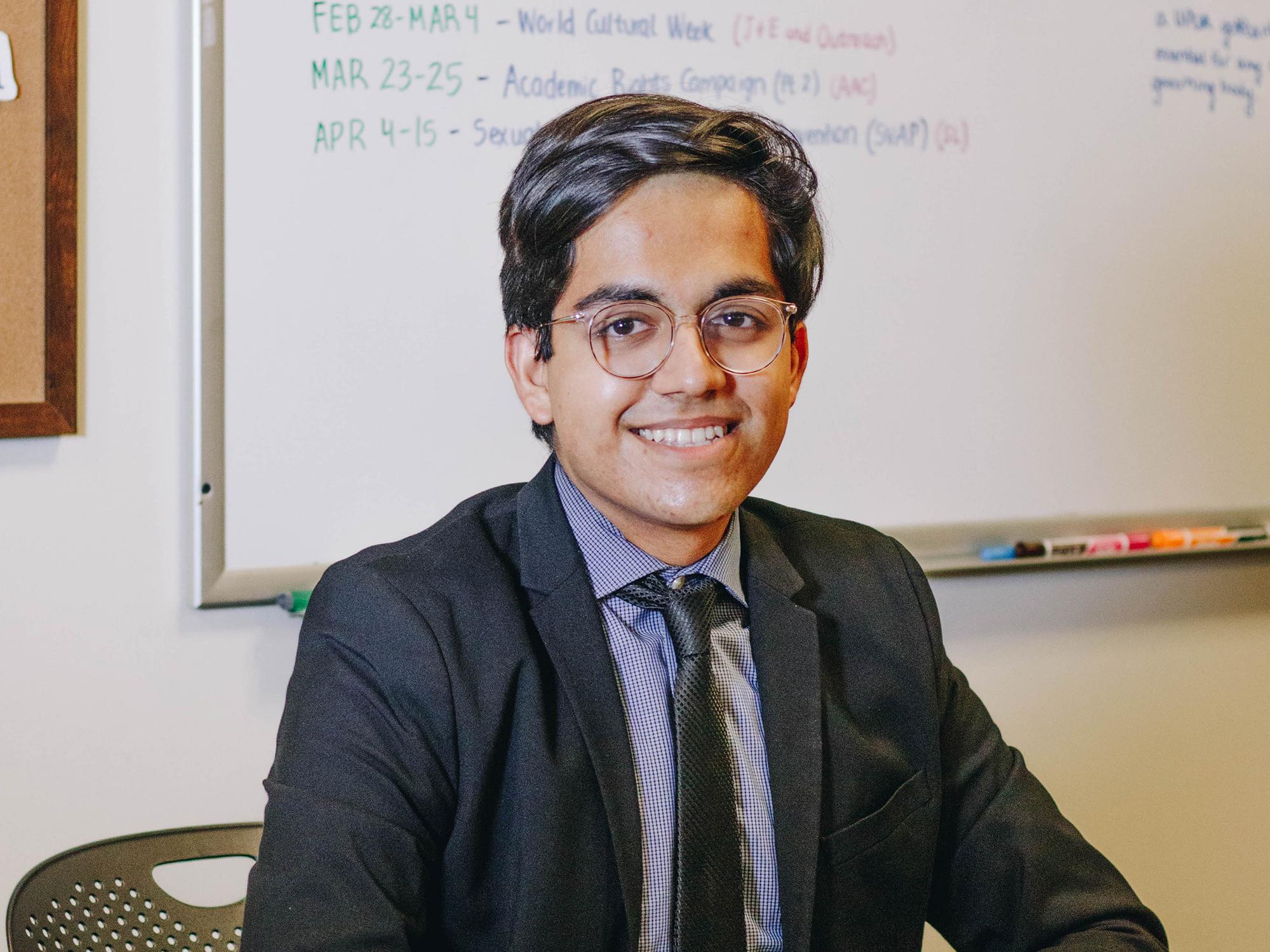 Taran Samarth, the spring 2023 College of the Liberal Arts student marshal, sits in front of a white board